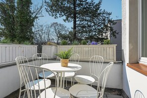 A patio with white metal chairs and a round table, surrounded by a white fence and greenery.