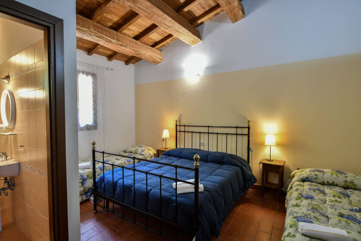 A bedroom with a metal bed frame, two bedside tables with lamps, a window with curtains, and a bathroom with a sink and mirror.