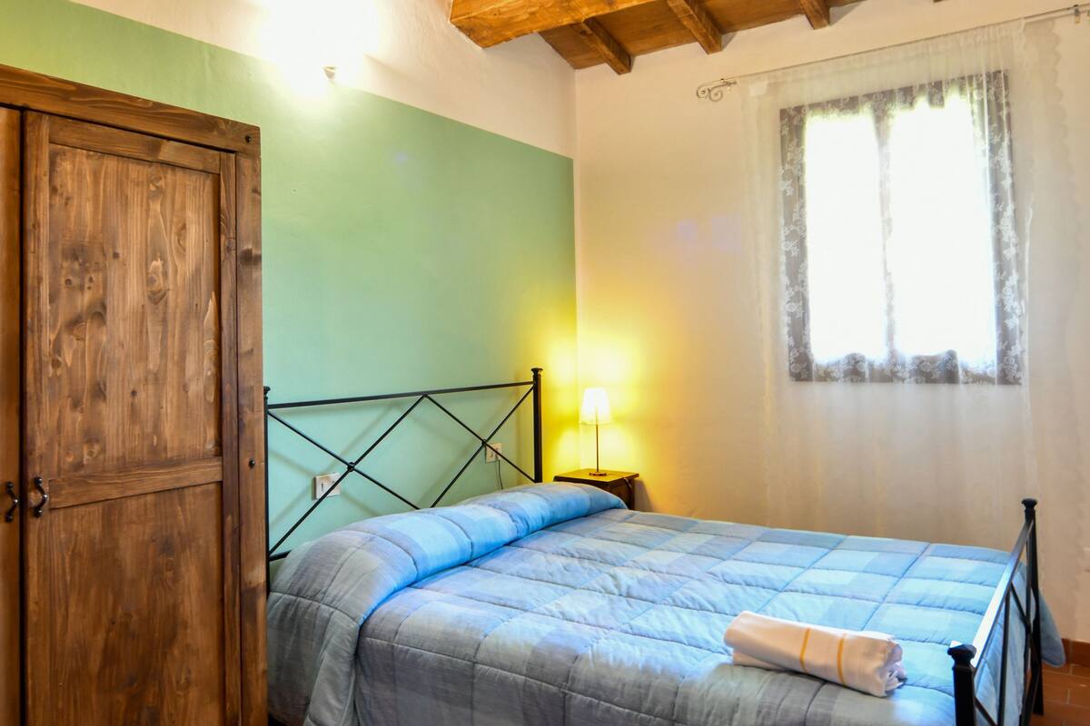 A bedroom with a wooden ceiling, a bed with a blue bedspread, a wooden wardrobe, and a window with a curtain.