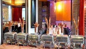 A restaurant with a modern interior, featuring a long dining table with multiple silver buffet stations, red curtains, and a warm ambient lighting. - Hotel Sharan (Rahata)
