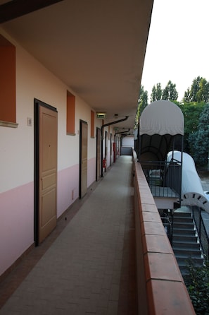 Un pasillo de hotel con puertas, una zona de estar exterior cubierta y una escalera que lleva a un nivel superior.