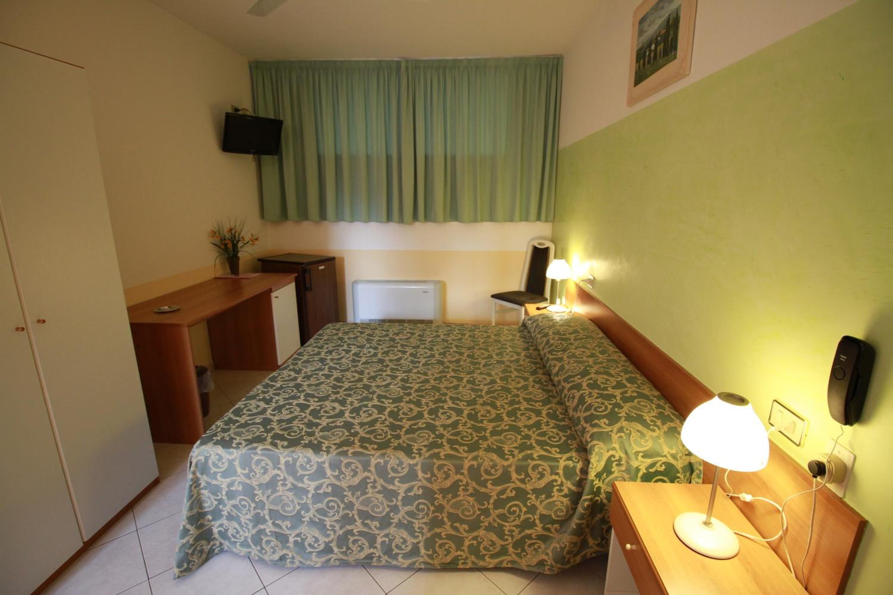 Ein Hotelzimmer mit einem Bett, einem Schreibtisch, einem Sessel, einer Lampe und einem Fernseher an der Wand.