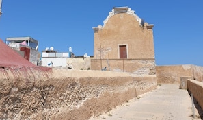 A building with a simple, unadorned facade and a small door, situated on a raised platform with a paved walkway leading to it. - Dar Statia 3 (El Jadida)
