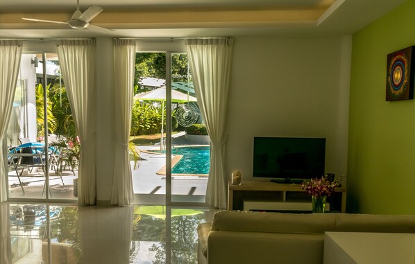 A modern living room with a large TV, a sofa, and a view of a swimming pool through glass doors.