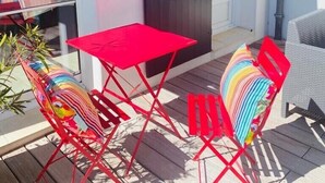 Un patio con una mesa y sillas rojas, una puerta oscura y una pared blanca con un ventanal circular.