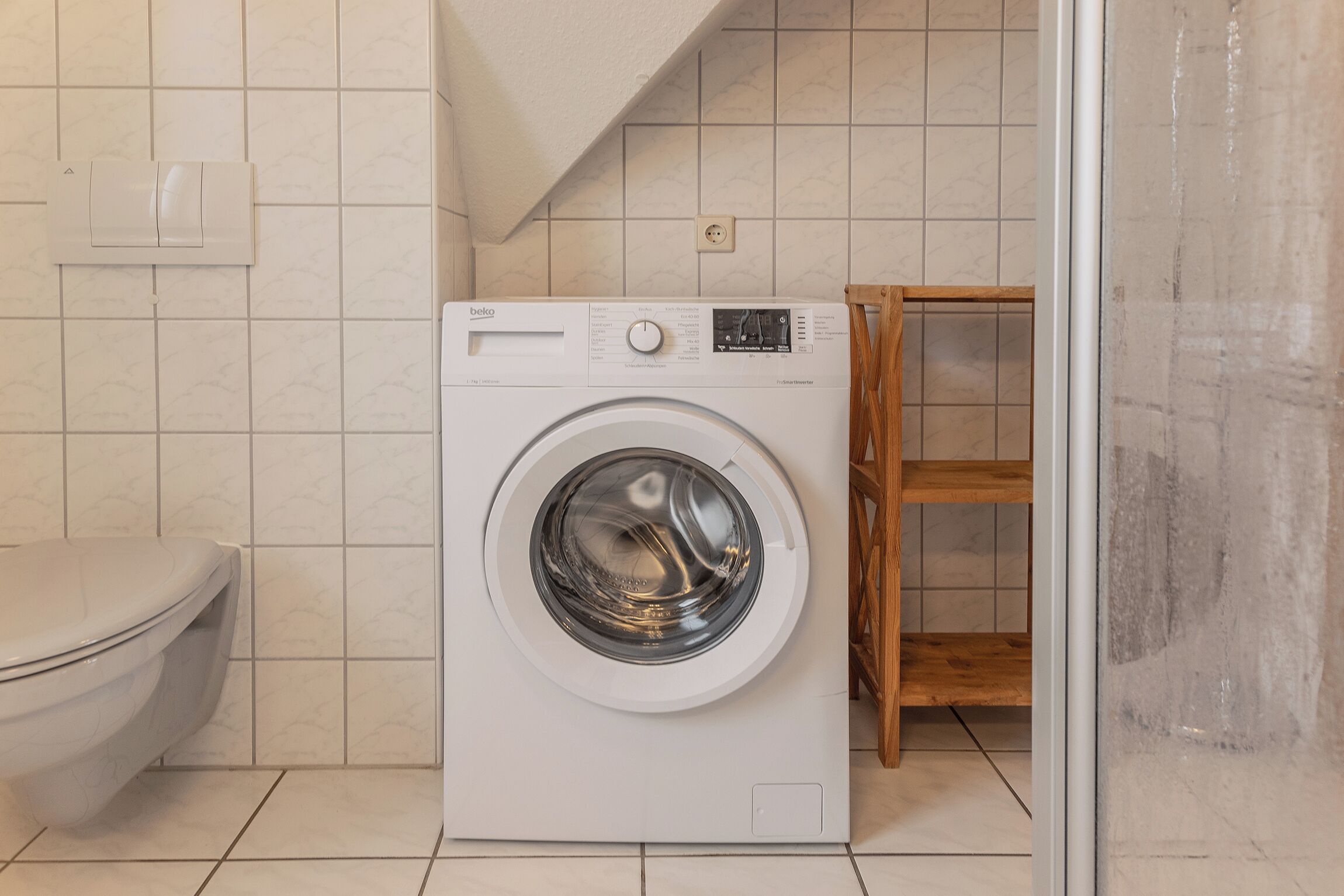 A compact laundry area with a front-loading washing machine, a toilet, and a wooden shelf unit.