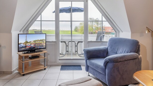 A living room with a television, a blue armchair, and a view of a balcony with a table and chairs.