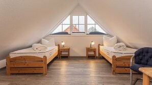A small, triangular attic bedroom with two wooden beds, a window with a view of a house, and a chair.