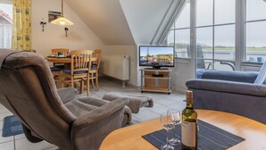 A cozy living room with a sofa, armchair, and dining table. A television is mounted on the wall, and a bottle of wine is on the table.