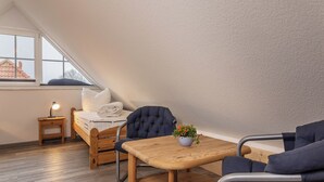 A cozy loft bedroom with a sloped ceiling, a wooden bed, a small bedside table with a lamp, a dark upholstered chair, and a wooden table with a potted plant.