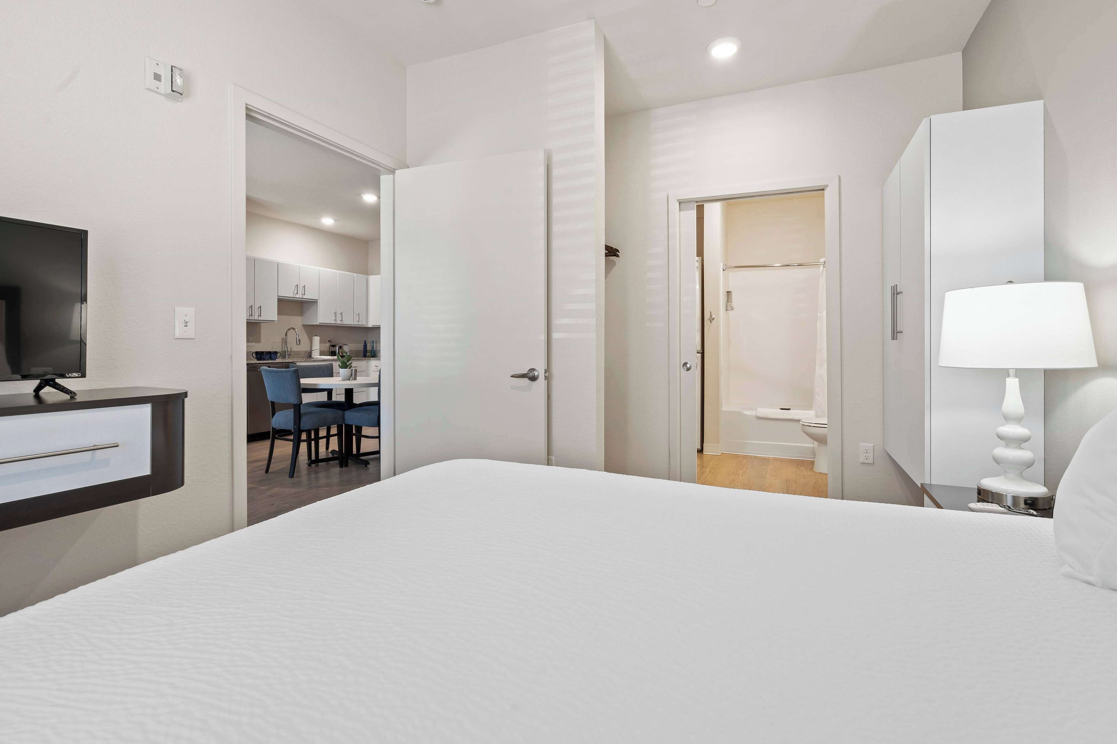 Una habitación de hotel moderna con una cama grande, un escritorio con televisor, un baño con ducha y un área de comedor visible a través de una puerta abierta.