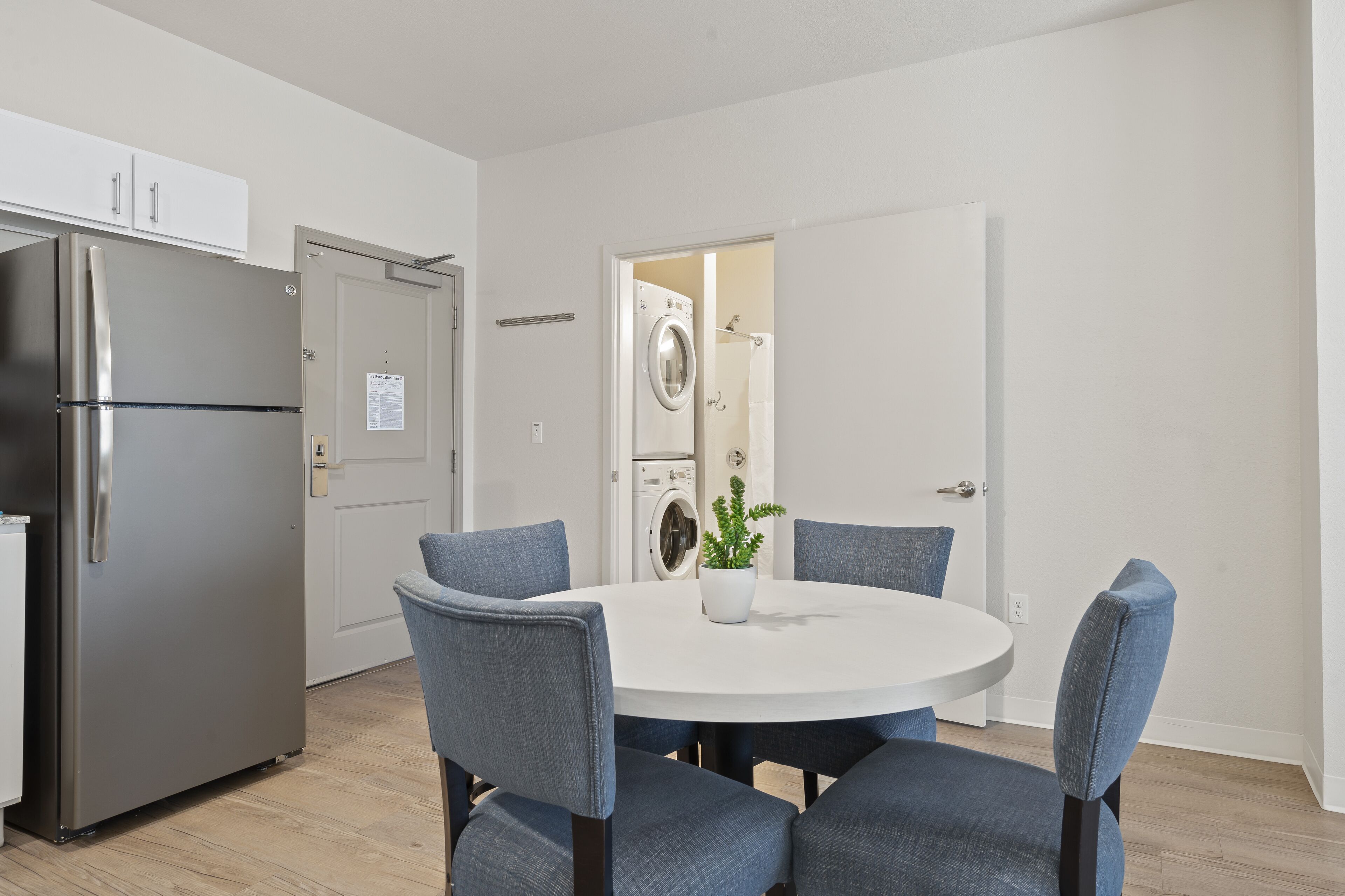 A modern kitchen with a stainless steel refrigerator, a dining table with four chairs, and a potted plant.