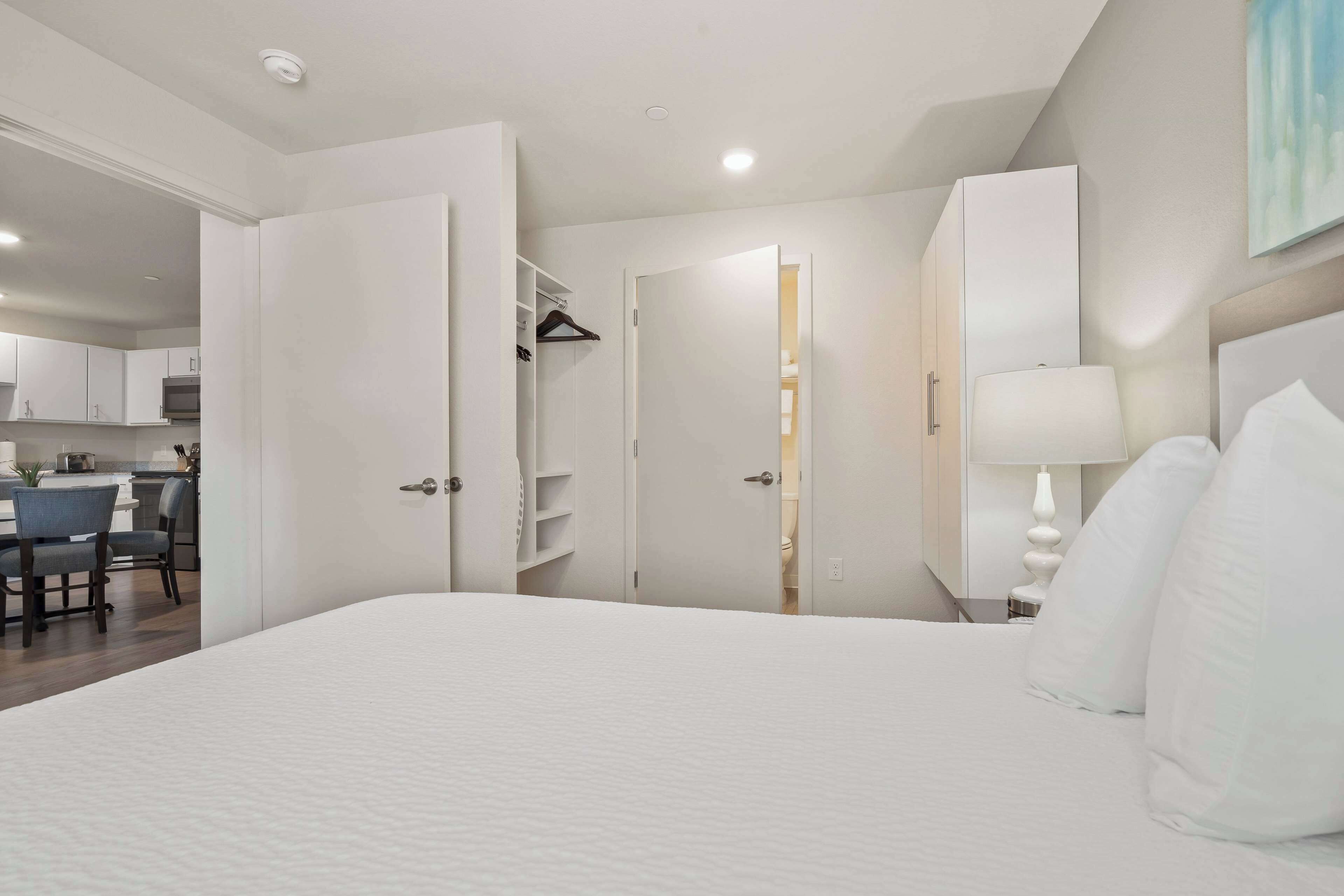 Habitación de hotel con una cama grande, una mesita de noche con lámpara, un armario y una puerta que da a un baño.
