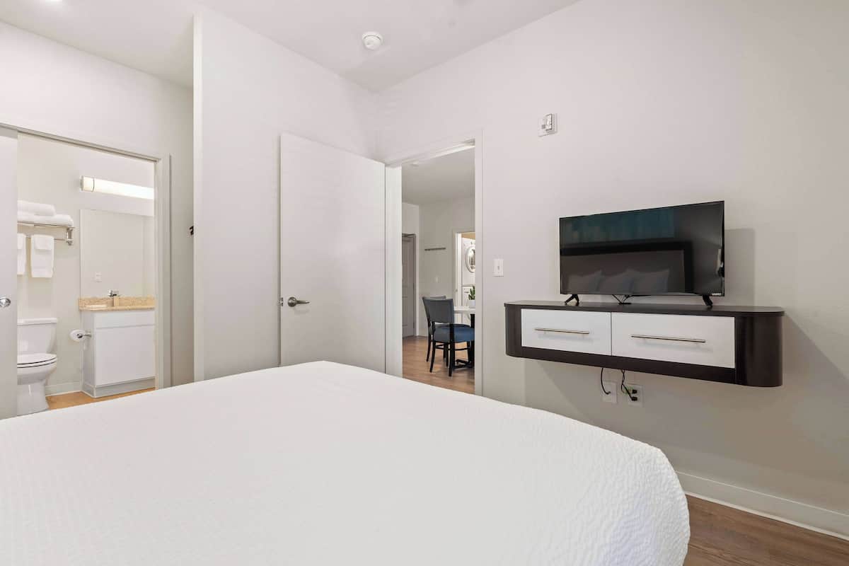 A hotel room with a large bed, a TV mounted on the wall, a bathroom with a toilet and sink, and a hallway leading to another room.