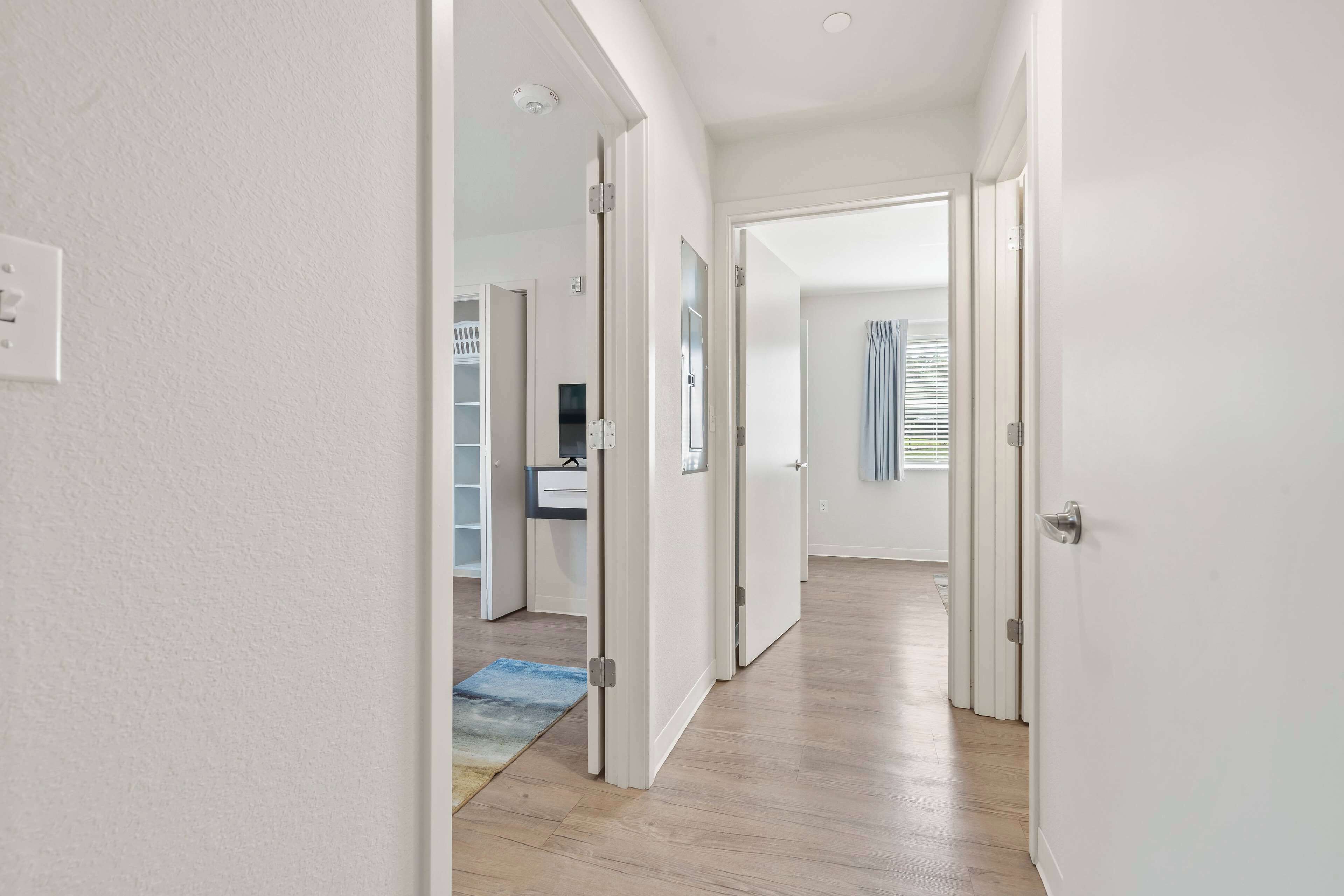 Un pasillo con paredes blancas, suelo de madera y una puerta que da a una habitación con un escritorio y una silla.