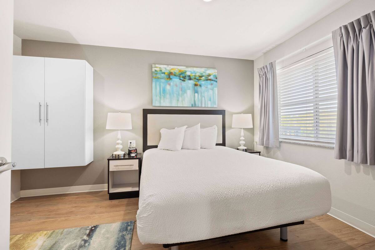 A modern bedroom with a large bed, a nightstand with a lamp, a wardrobe, and a painting on the wall.