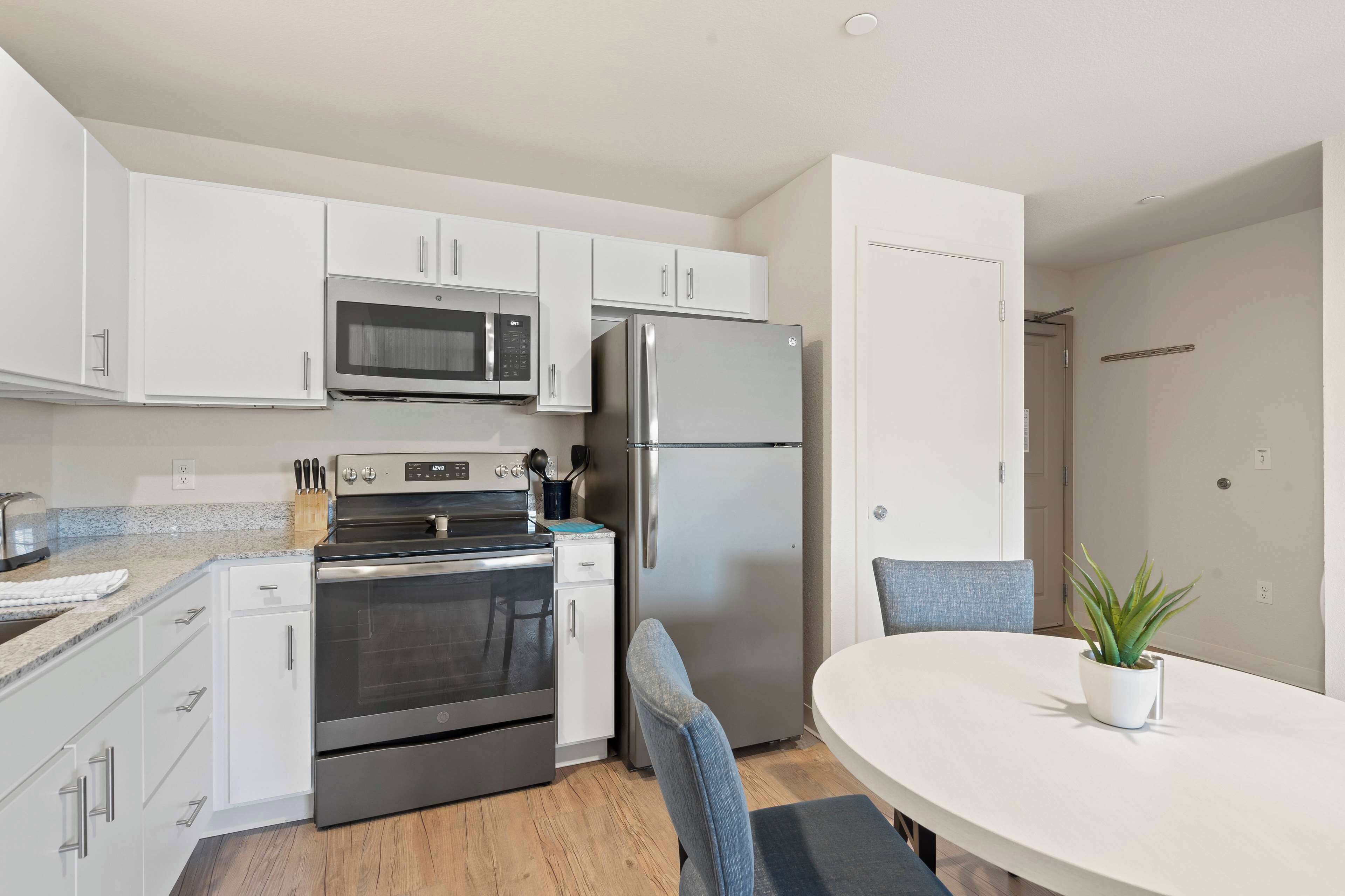 A modern kitchen with white cabinets, a stainless steel refrigerator, an oven, and a microwave. There is a dining table with a potted plant.