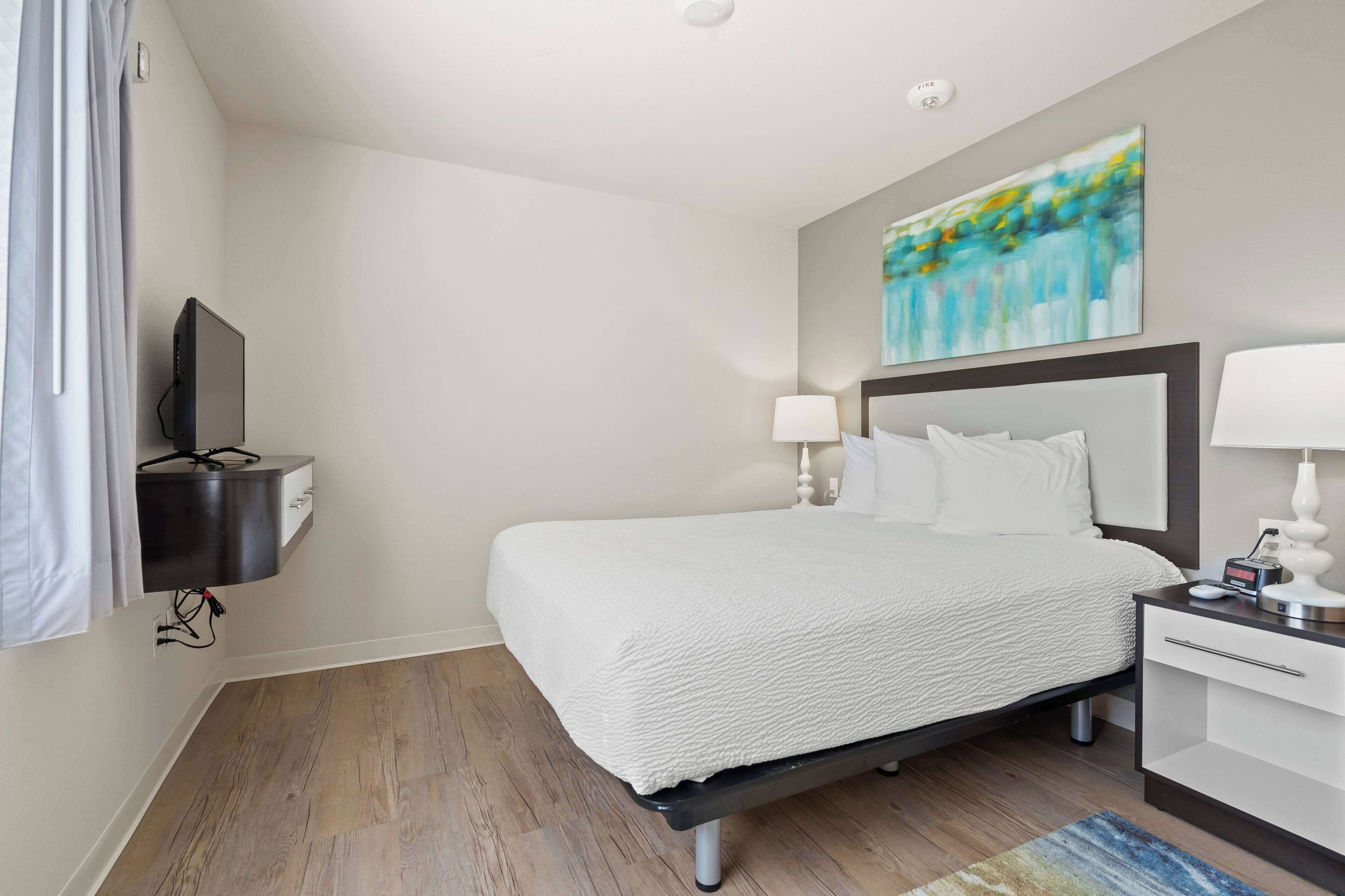 A hotel room with a bed, bedside table, lamp, and a painting on the wall.