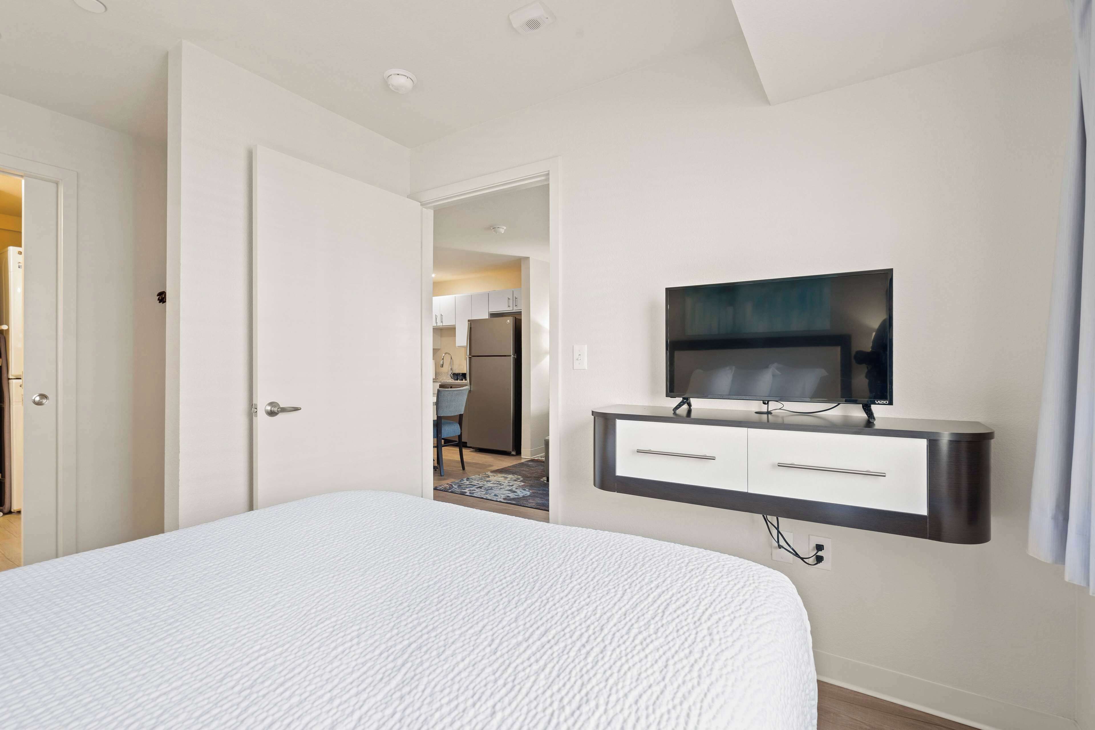 A modern hotel room with a large bed, a mounted TV, and a view into the kitchen area.
