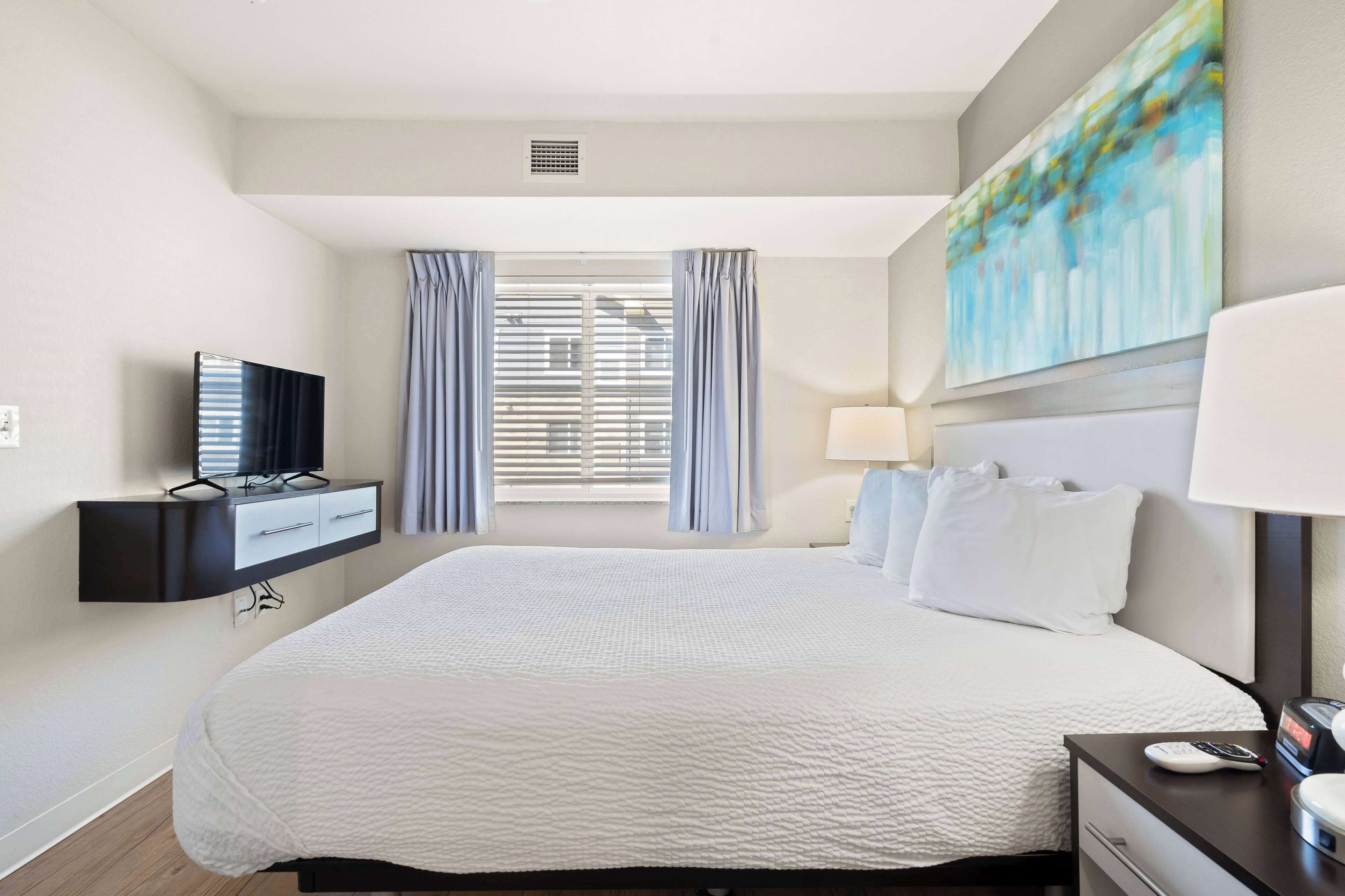 Habitación de hotel con una cama grande, un televisor sobre un mueble negro, un escritorio con una lámpara y un cuadro en la pared.