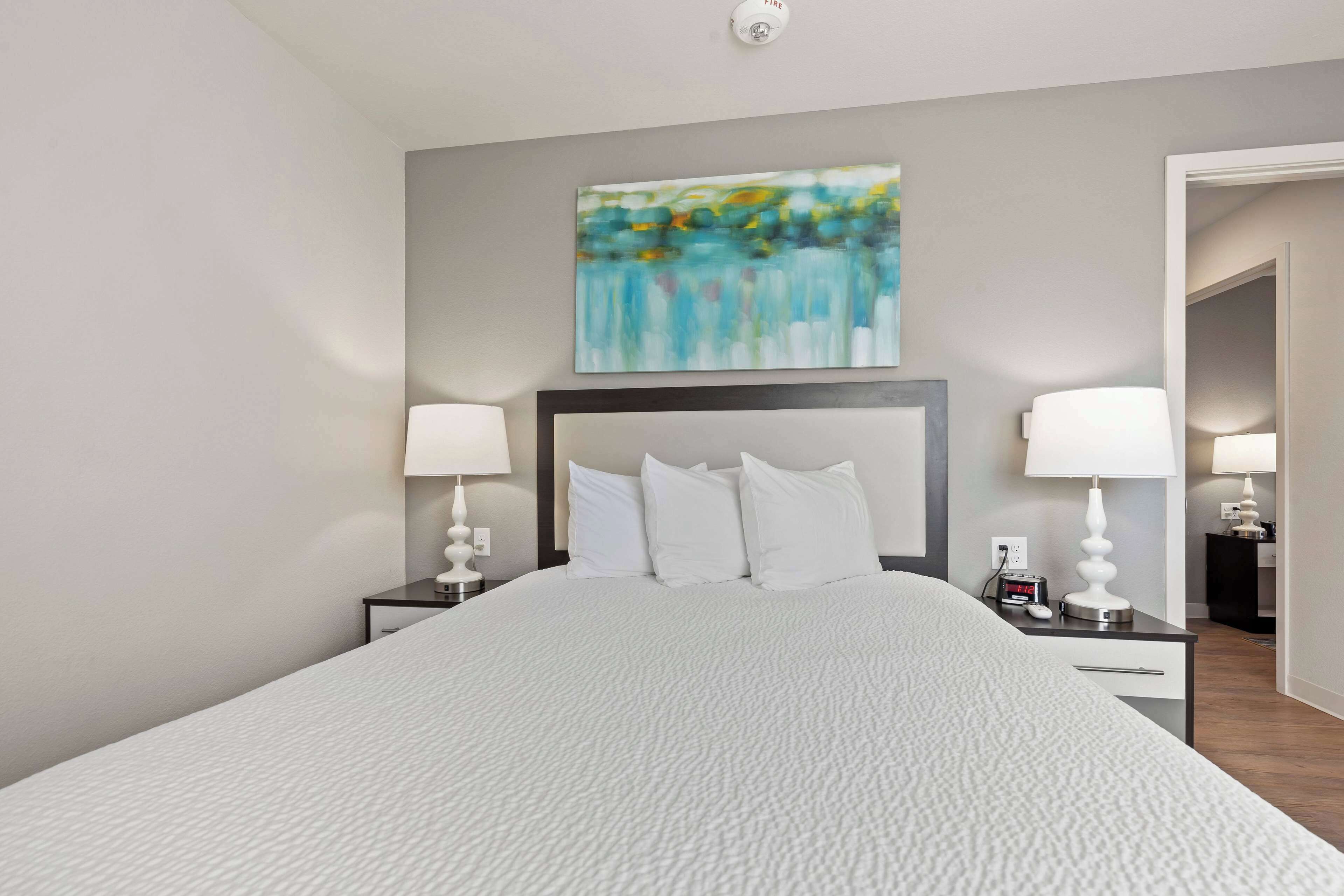 A neatly made bed with a white comforter, two bedside tables with lamps, and a modern abstract painting above the bed.