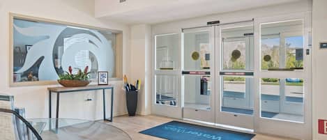 Un espace d’accueil moderne avec un bureau en verre, un paillasson et une grande fenêtre donnant sur l’extérieur.