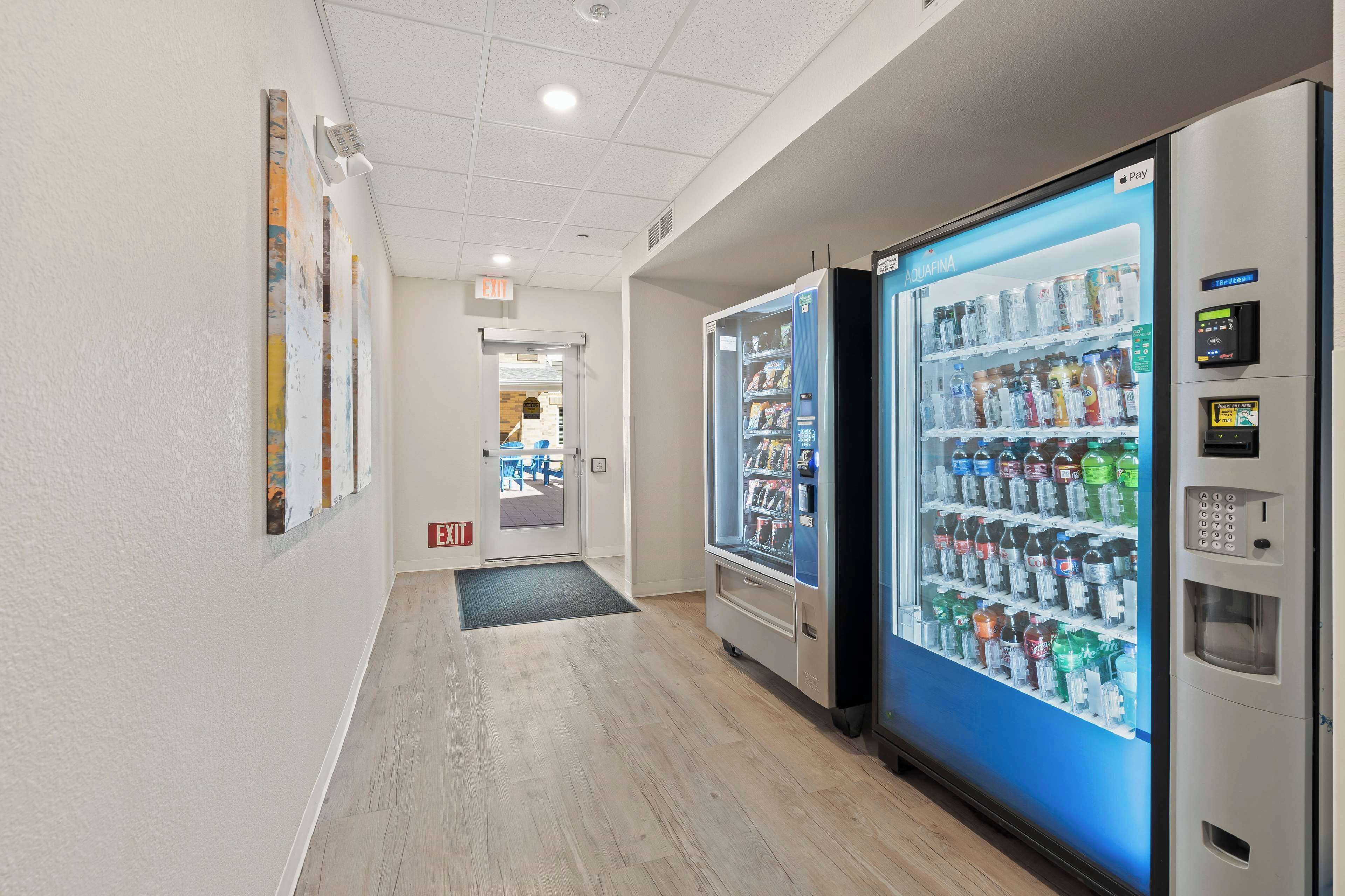 A hallway with two vending machines stocked with beverages and snacks, a door leading to an outdoor area, and a wall with abstract art.