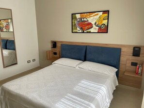 Camera d'albergo con un letto, una testiera, un comodino, uno specchio e un quadro raffigurante automobili sulla parete.