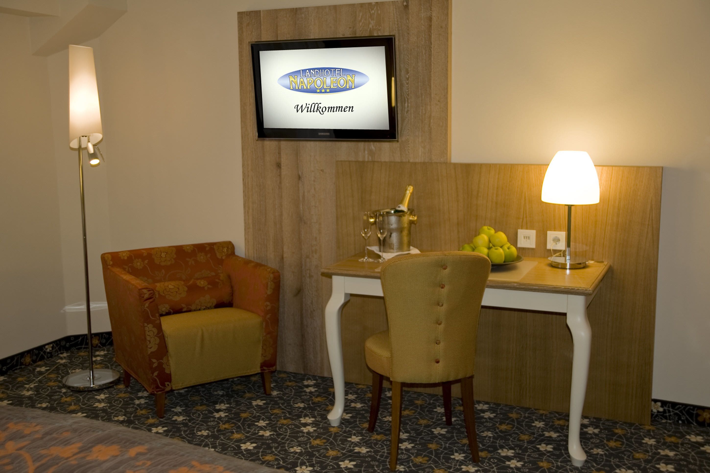 A hotel room with a television displaying "Landhotel Napoleon Willkommen," a small table with a bottle and glasses, a chair, and a lamp.