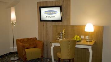 A hotel room with a television displaying "Landhotel Napoleon Willkommen," a small table with a bottle and glasses, a chair, and a lamp.
