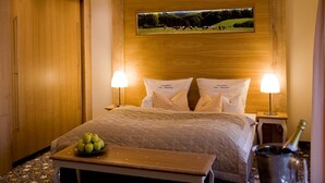 A hotel room with a large bed, two bedside lamps, a wooden headboard, a framed picture above the bed, a bedside table with a bowl of apples, and a bottle of champagne. - Landhotel Napoleon (Wipperfuerth)