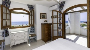 A bedroom with a view of the sea, a ceiling fan, a dresser, a chair, and a wooden cabinet.