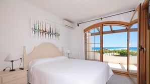 A bedroom with a large bed, a nightstand, a lamp, a wall-mounted air conditioner, and a window with a view of the beach.
