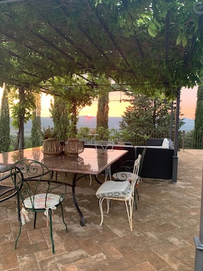 Um pátio externo com mesa e cadeiras, churrasqueira e vista para o pôr do sol nas montanhas.