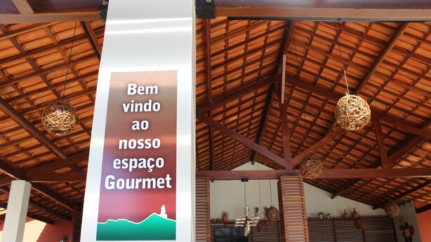 A sign with the text "Bem vindo ao nosso espaço Gourmet" and a view of a covered outdoor area with a wooden ceiling and hanging light fixtures.