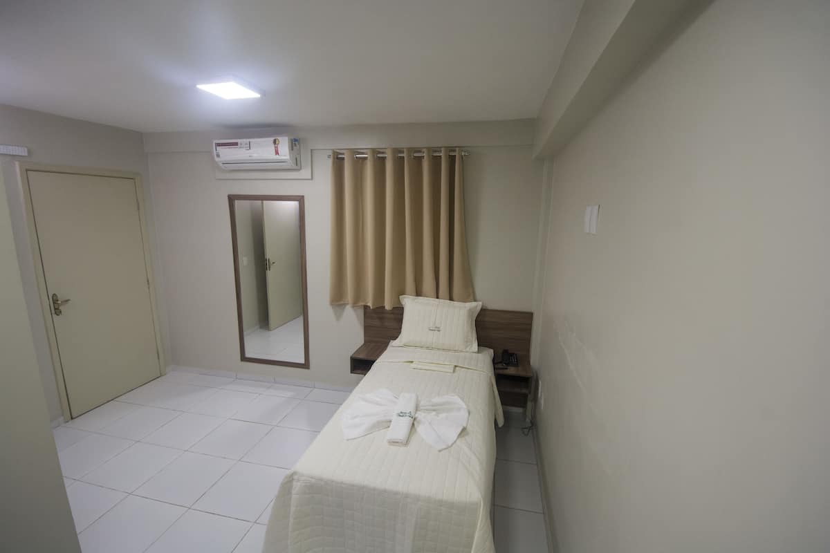 Une chambre d’hôtel avec un lit simple, une table de chevet, un miroir, une porte et une fenêtre avec des rideaux.