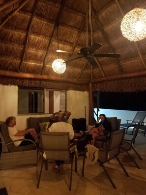 Un espace extérieur aménagé avec un toit de chaume, doté d’un ventilateur de plafond et de luminaires suspendus, entouré de fauteuils et d’un canapé.
