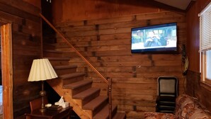 Een houten interieur met een trap, een aan de muur bevestigde televisie, een tafel met een lamp en een gestreept bankstel.