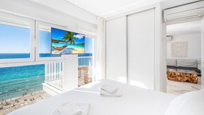 A hotel room with a large window overlooking a beach, a bed with white linens, and a television displaying a beach scene. - Veracruz 11-B (Benidorm)