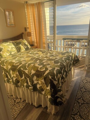 A bedroom with a bed, curtains, a view of the ocean, and a balcony with chairs.
