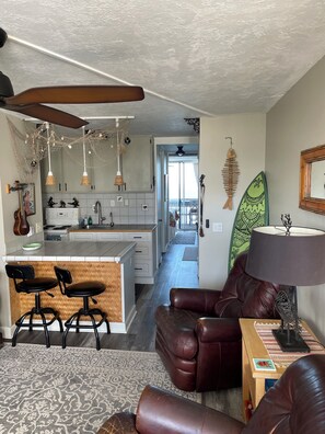 A modern kitchen with pendant lights, a dining area with leather chairs, a surfboard decoration, and a view to a balcony.