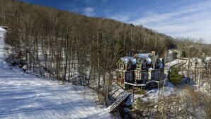A large, multi-story house with a prominent front porch and a wrap-around deck, surrounded by a snowy landscape and dense forest. - Luxury 6br Solitude Ge- Indoor Bball Court, Private Tub 6 Bedroom Home by RedAwning (Ludlow)