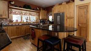A kitchen with wooden cabinets, a stainless steel refrigerator, a large island with stools, and a window with a floral curtain. - Luxury 6br Solitude Ge- Indoor Bball Court, Private Tub 6 Bedroom Home by RedAwning (Ludlow)