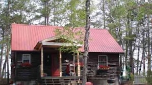A rustic cabin with a red roof, a porch with a red chair, and a fire pit.