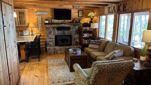 A cozy cabin interior with a fireplace, wooden walls, a sofa, a patterned armchair, and a bookshelf.