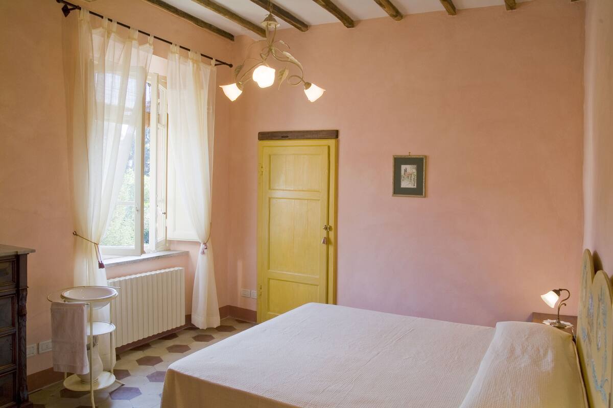 Een slaapkamer met een bed, een raam met gordijnen, een radiator, een houten deur en een nachtkastje met een lamp.