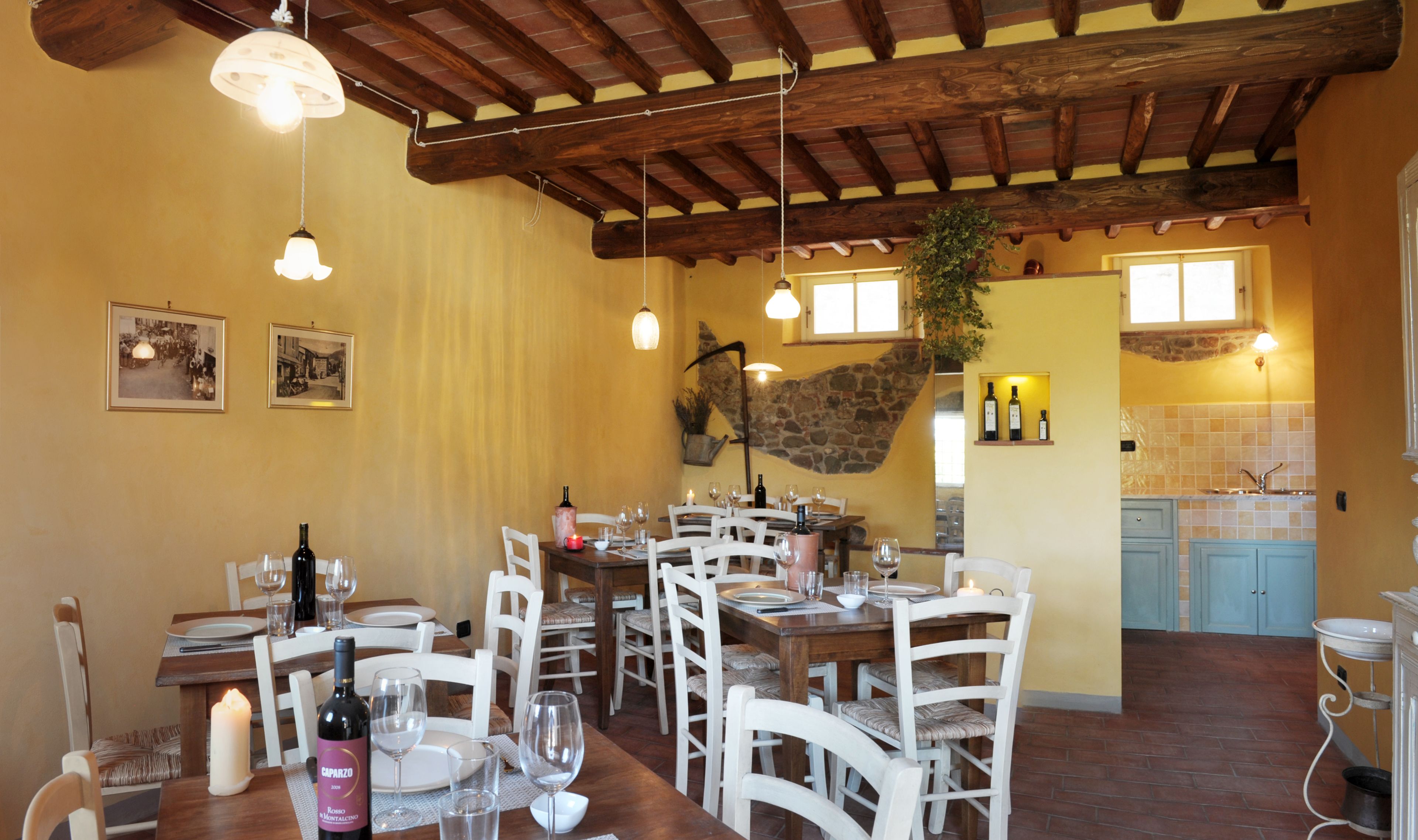 A dining area with wooden tables and chairs, wine glasses, and bottles. There are framed pictures on the wall and a stone wall in the background.