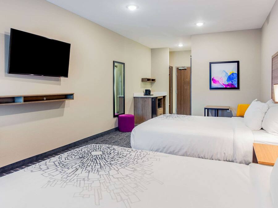 A hotel room with two beds, a flat-screen TV mounted on the wall, a small desk with a lamp, and a colorful abstract painting on the wall.