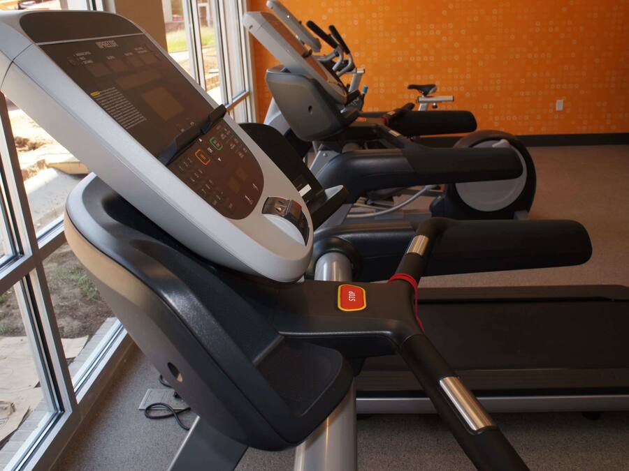 Una fila de bicicletas estáticas, con pantallas digitales, ubicadas en una habitación con pared naranja y ventanas amplias.