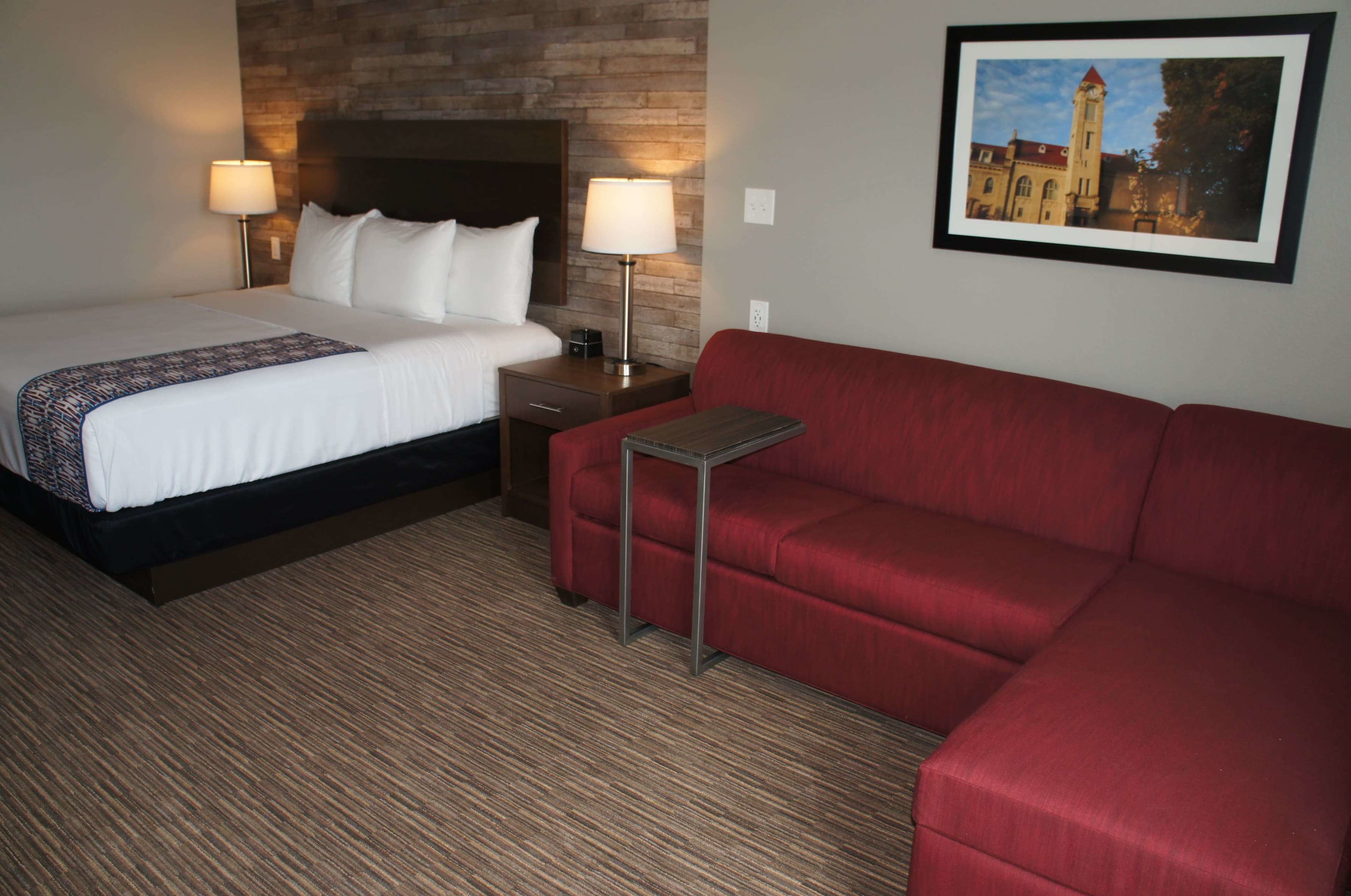A hotel room with a bed, a red sofa, a nightstand with a lamp, and a framed picture on the wall.